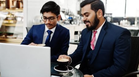 Two indian business man in suits sitting at office on cafe, looking at laptop and drinking coffee.